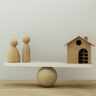 Wooden people figures and a small house balanced on a seesaw, symbolizing the delicate balance and control needed to manage family assets and secure family property