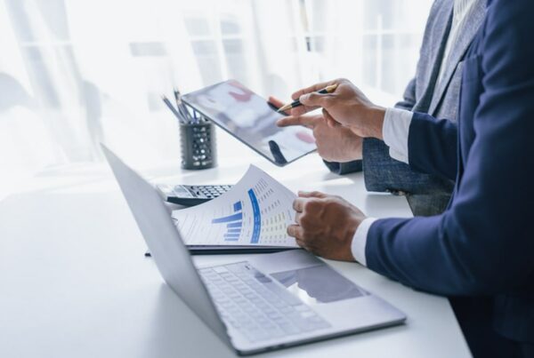 Two professionals discuss a report with financial graphs while working on a laptop and tablet in an office setting. The image symbolizes how AV does the heavy lifting, allowing family principals to focus on decision making